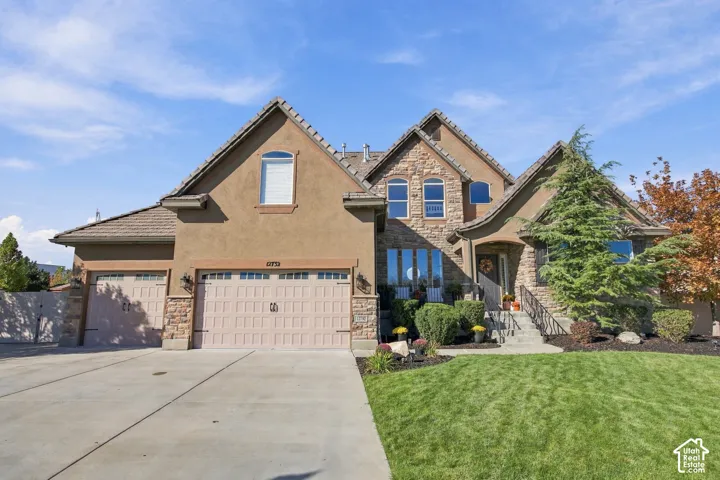 Stucco and stone exterior with a multi-car driveway, attached garages, a manicured lawn, and mature landscaping