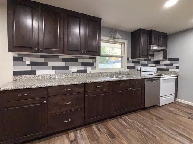 Dark wood-finish cabinetry, light speckled countertops, multi-tone backsplash, wood-finish flooring, and a double basin sink
