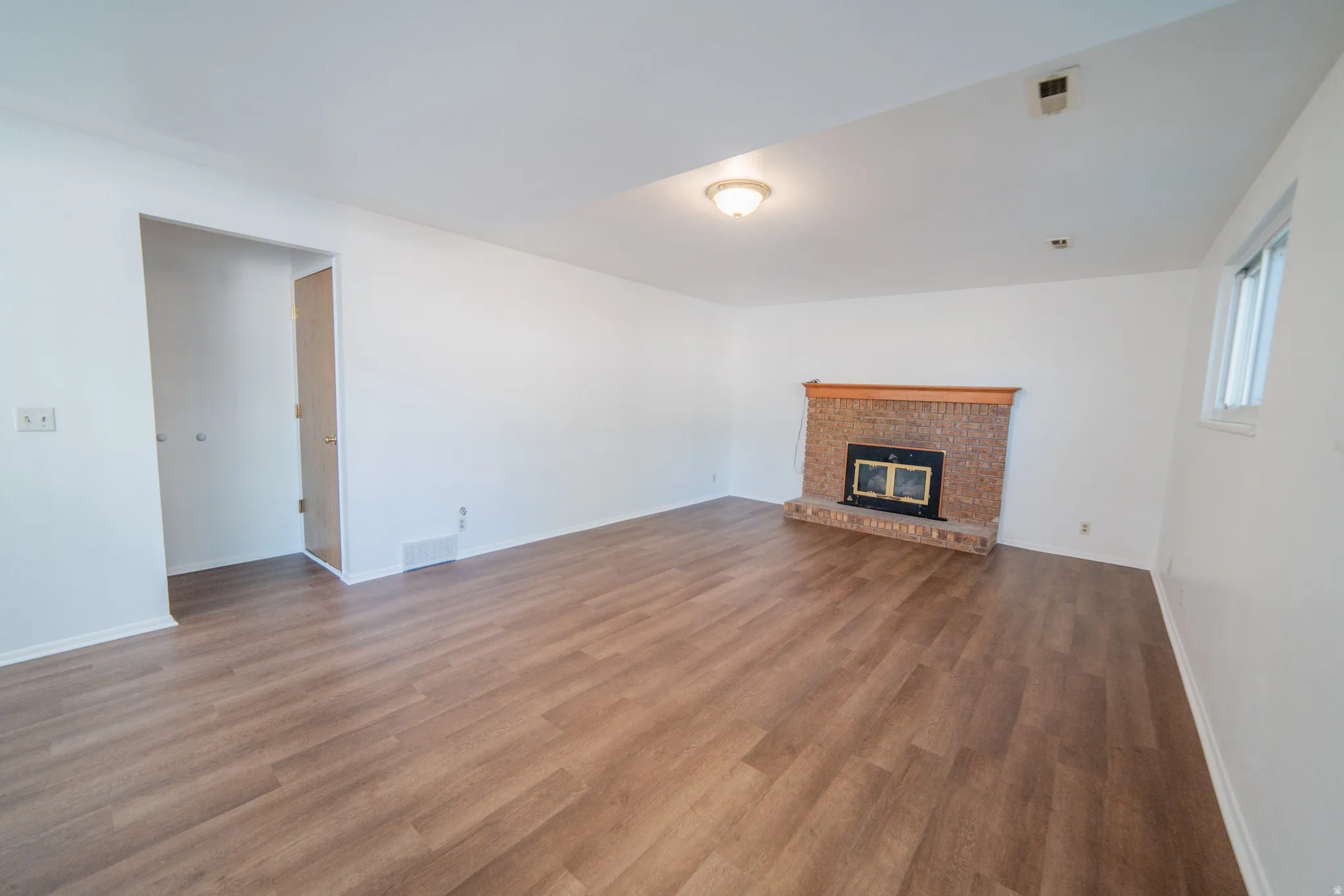 Wood-finish flooring, brick-faced fireplace with wood mantel, white walls, and a window providing natural light