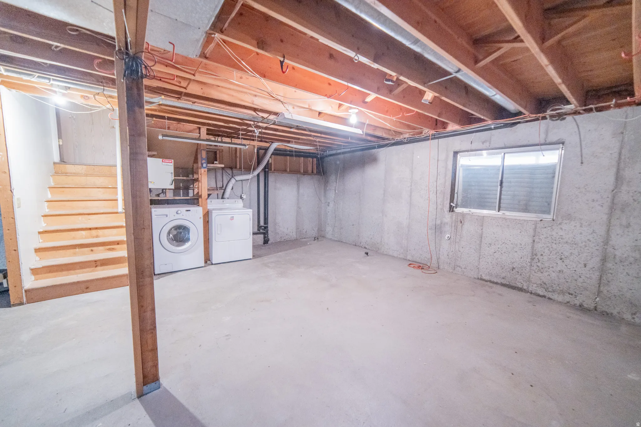 Unfinished basement featuring exposed wood joists, concrete walls, a single-pane window, and a utility area with laundry appliances