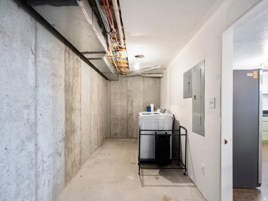 Unfinished basement with concrete walls and exposed overhead utilities, featuring electrical panels and a dedicated laundry area for ADU.