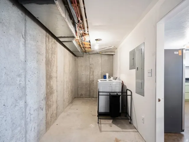 Unfinished basement with concrete walls and exposed overhead utilities, featuring electrical panels and a dedicated laundry area for ADU.