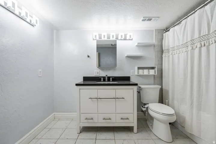 White vanity with dark countertop, square tile flooring, wall-mounted shelving, and recessed lighting