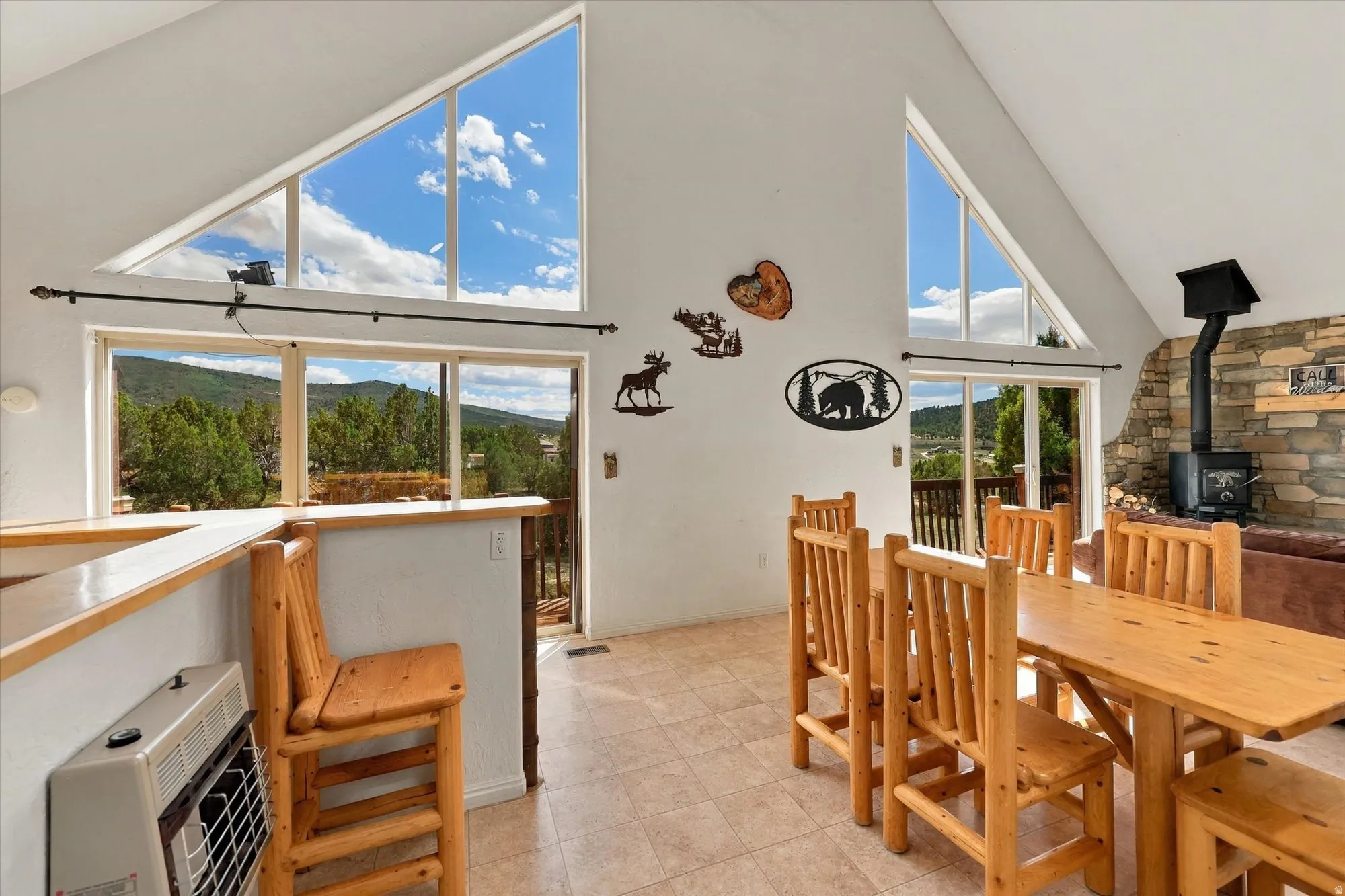 Vaulted ceiling with triangular windows, stone-clad fireplace with a wood-burning stove, large sliding glass doors, and light-toned tile flooring