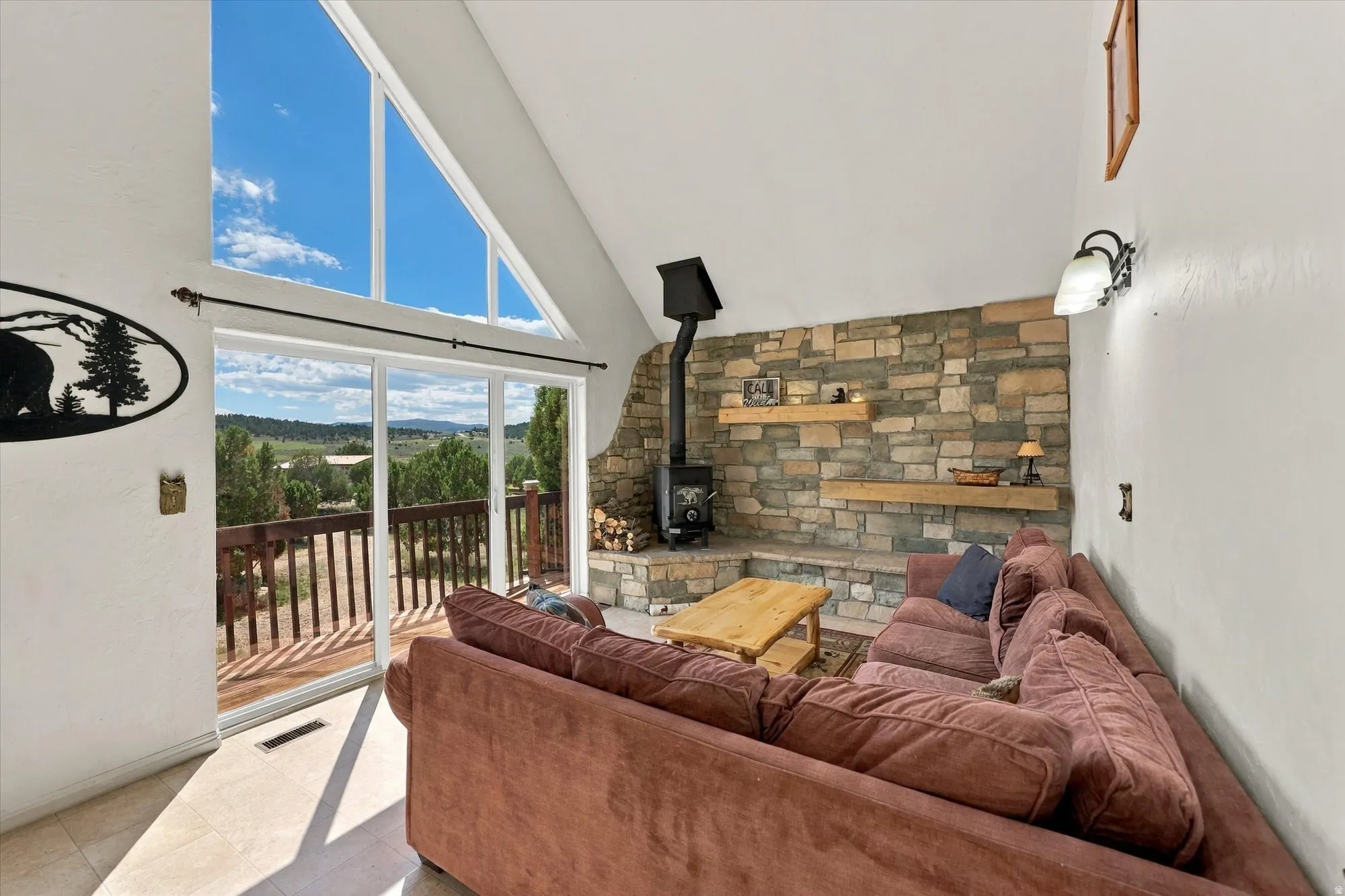 Vaulted ceiling with triangular window array, full-height stone accent wall, wood-burning stove with stone hearth, and sliding glass doors opening to an exterior deck