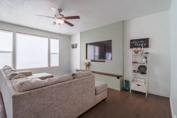 Wood-finish flooring throughout, ceiling fan with integrated lighting, multiple windows with blinds, and a built-in entertainment wall with a floating shelf