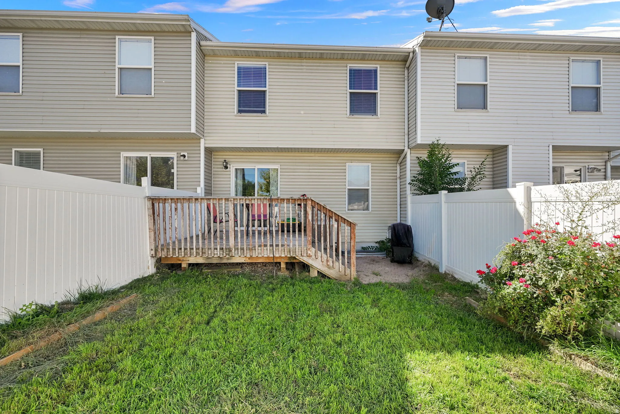 Wood deck with railing, white vinyl fencing, sliding glass door access, light-colored siding, and a landscaped shrub