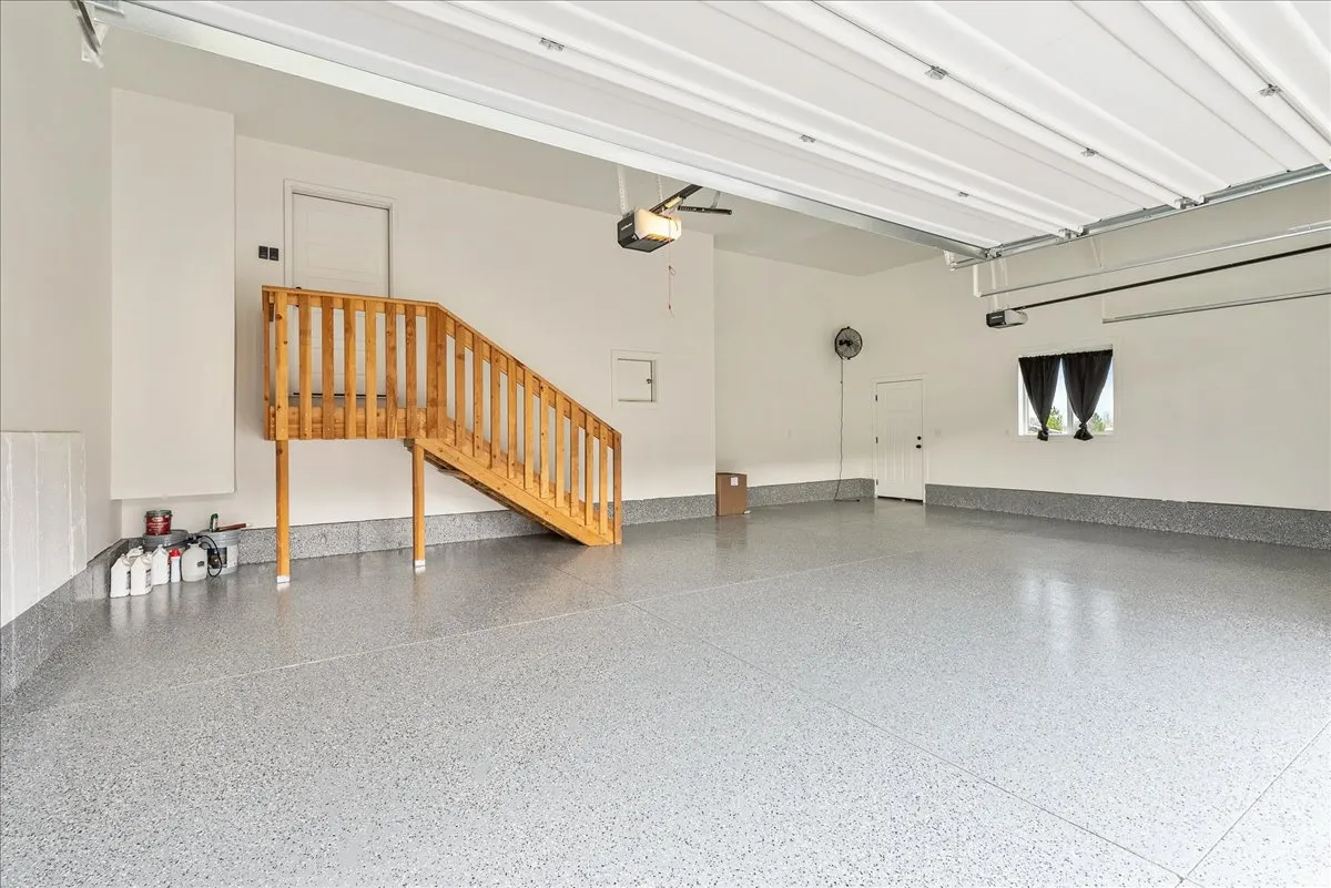 Spacious garage with light-colored, speckled flooring, overhead garage door system, and a natural wood staircase