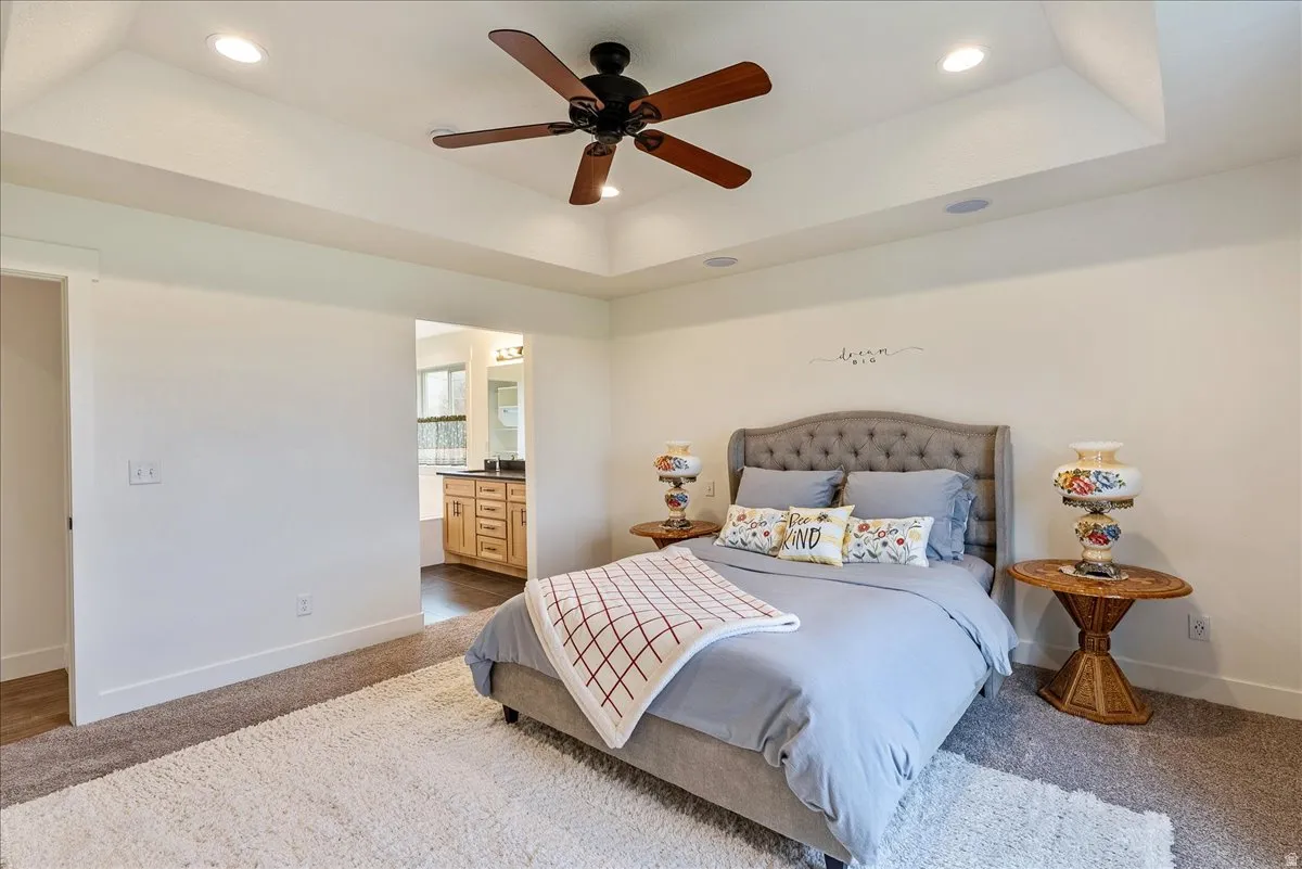 Recessed lighting, ceiling fan, tray ceiling, carpeted flooring, and a bathroom with a wood-finish vanity