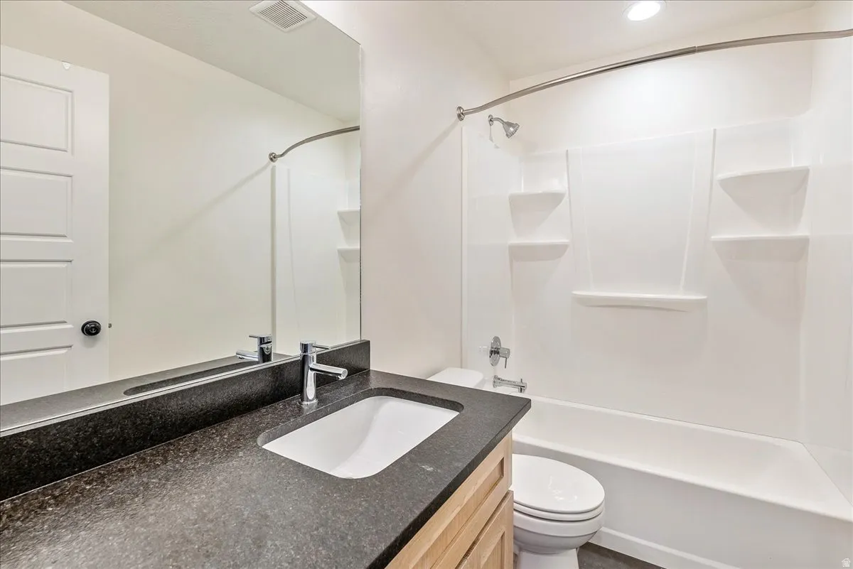 Bathroom featuring a full-width vanity mirror, dark countertop with an integrated sink, chrome faucet, wood-finish vanity cabinetry, and a white tub/shower combination with built-in shelving