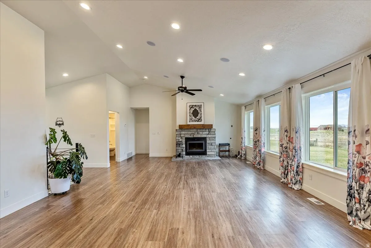 Vaulted ceilings with recessed lighting, wood-finish flooring, stone fireplace with wood mantel, ceiling fan, and multiple large windows
