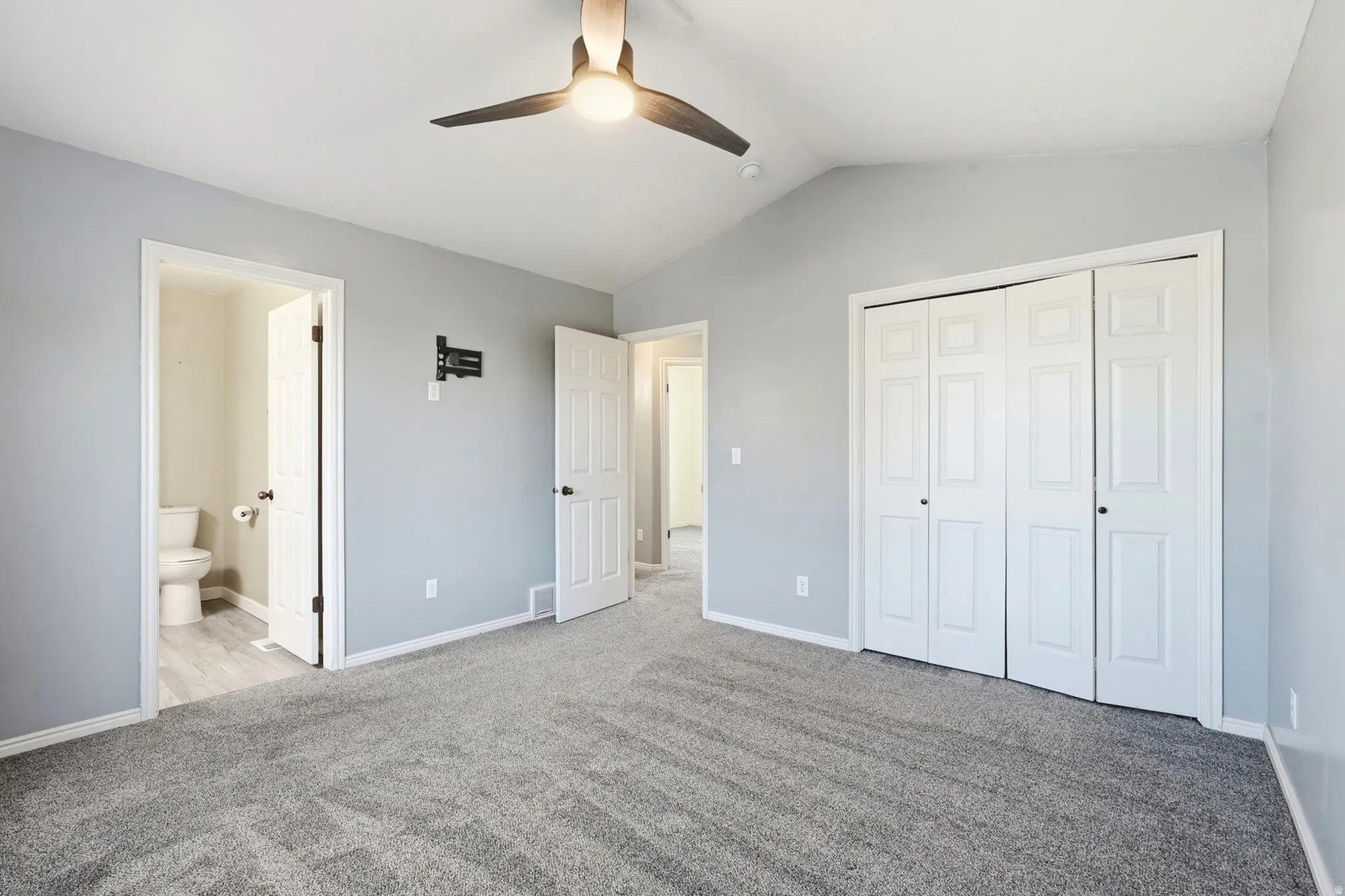 Vaulted ceiling with a ceiling fan, plush gray carpeting, light gray wall paint, white bi-fold closet doors, and a wall-mounted TV bracket