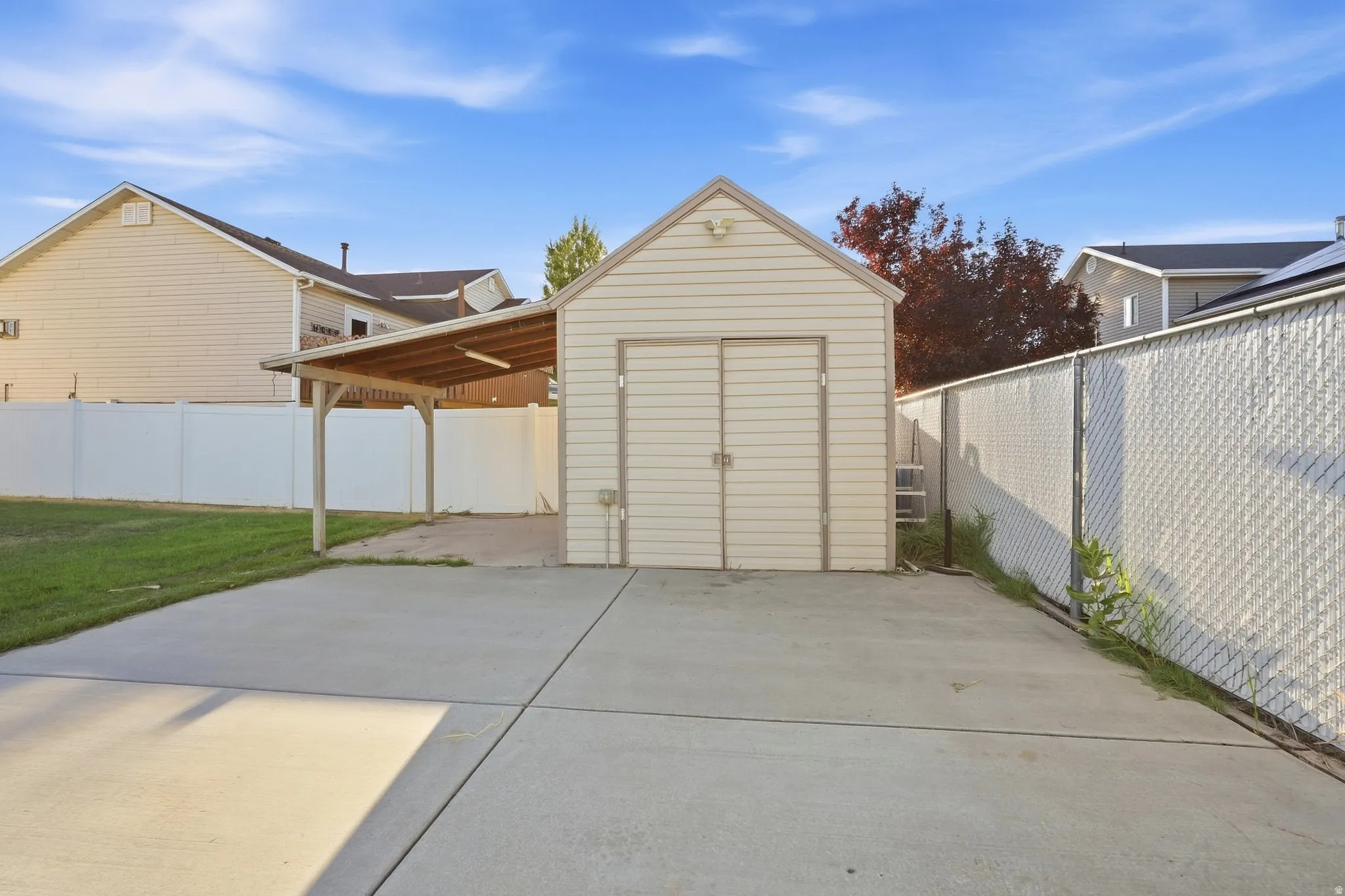 Detached storage shed with double doors, covered carport, concrete paving, and a chain-link fence
