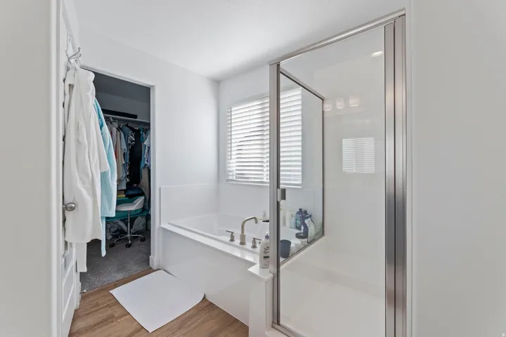 Walk-in shower with glass enclosure, soaking tub with deck-mounted fixtures, window with blinds, wood-finish flooring, and an open closet with shelving