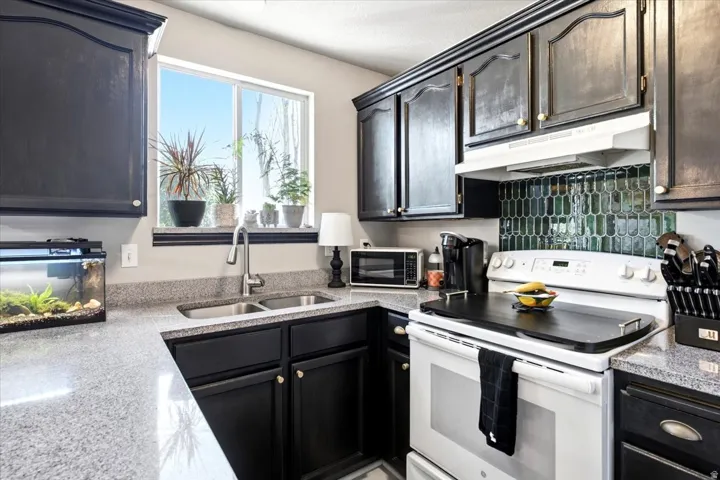 Kitchen featuring dark cabinetry, light-toned granite countertops, a stainless steel double basin sink, a window, and a green tile backsplash