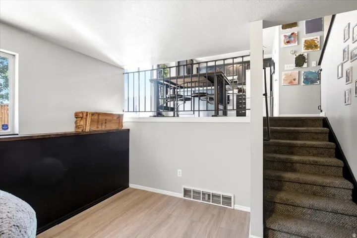 Wood-finish flooring, carpeted staircase with dark handrail, interior window with black metal railing, and white trim throughout