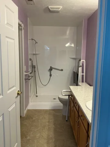 Walk-in shower with grab bars, single vanity with light-toned countertop, wood-finish cabinetry, and neutral-toned flooring