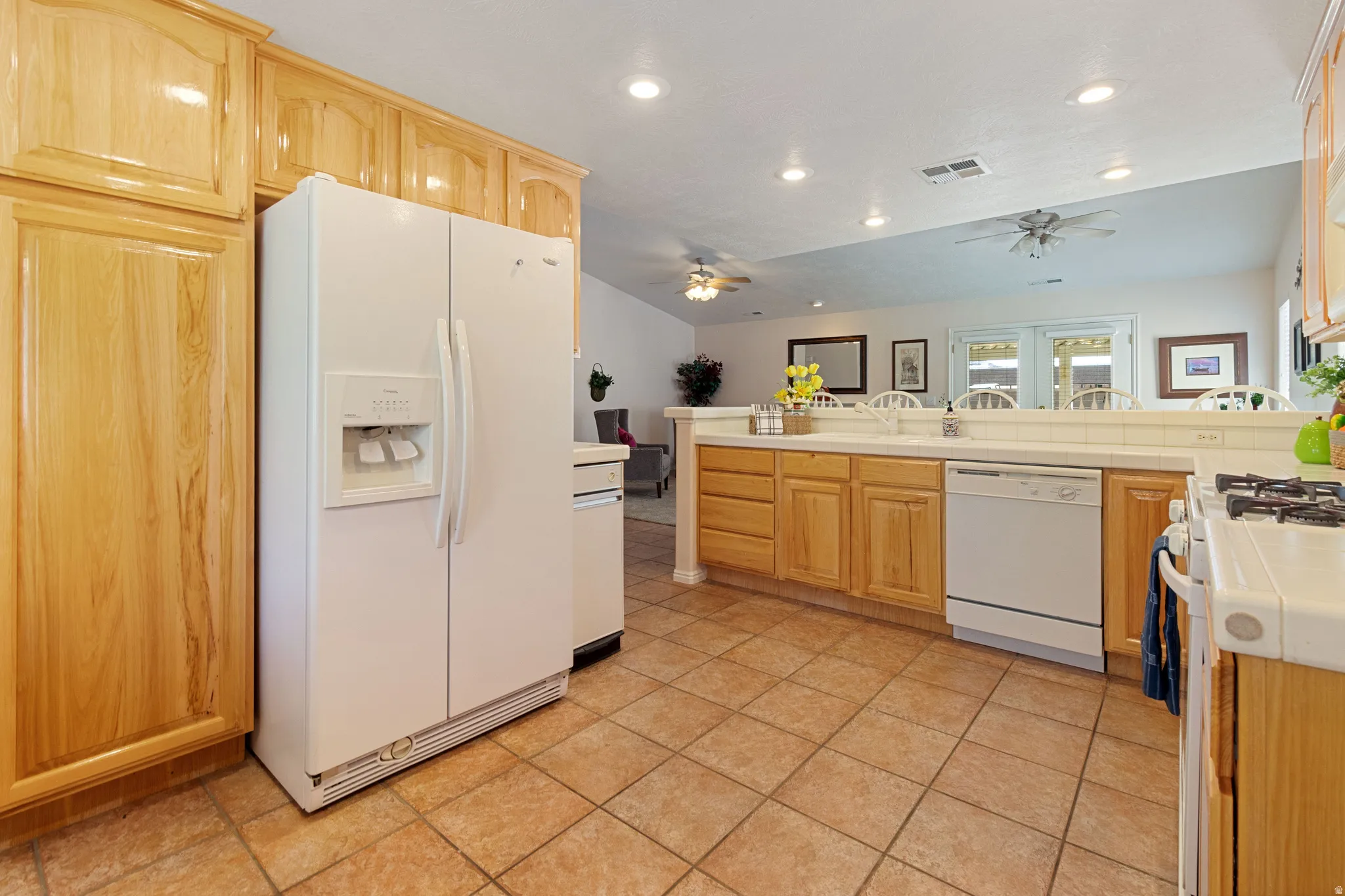 Kitchen featuring wood cabinetry, white appliances, tile flooring, recessed lighting, and a breakfast bar with a tile counter