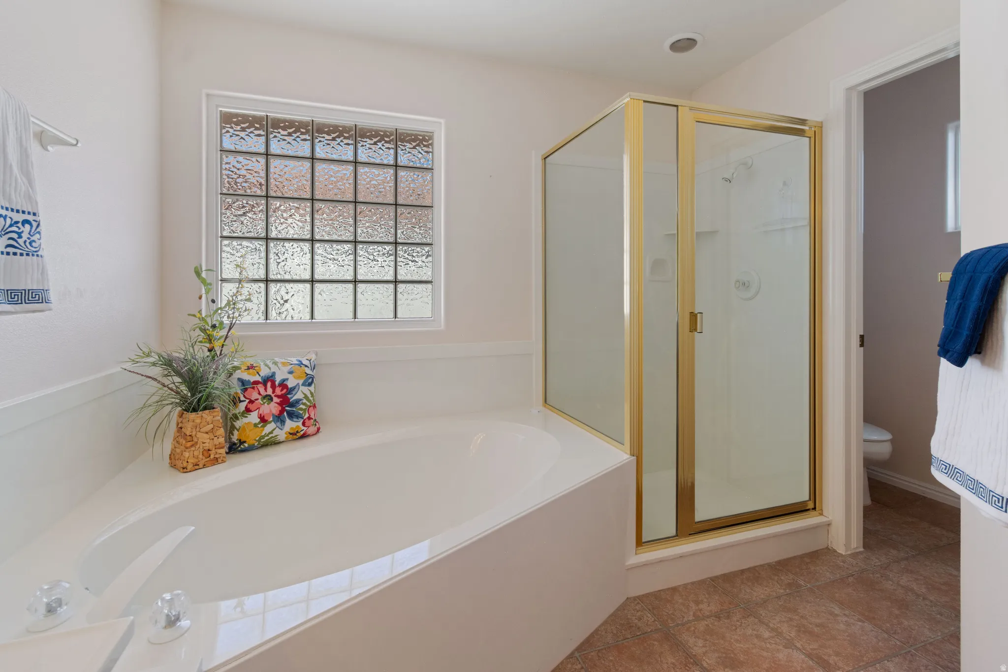 Corner soaking tub, glass block window, enclosed shower with gold-tone frame, and tile flooring