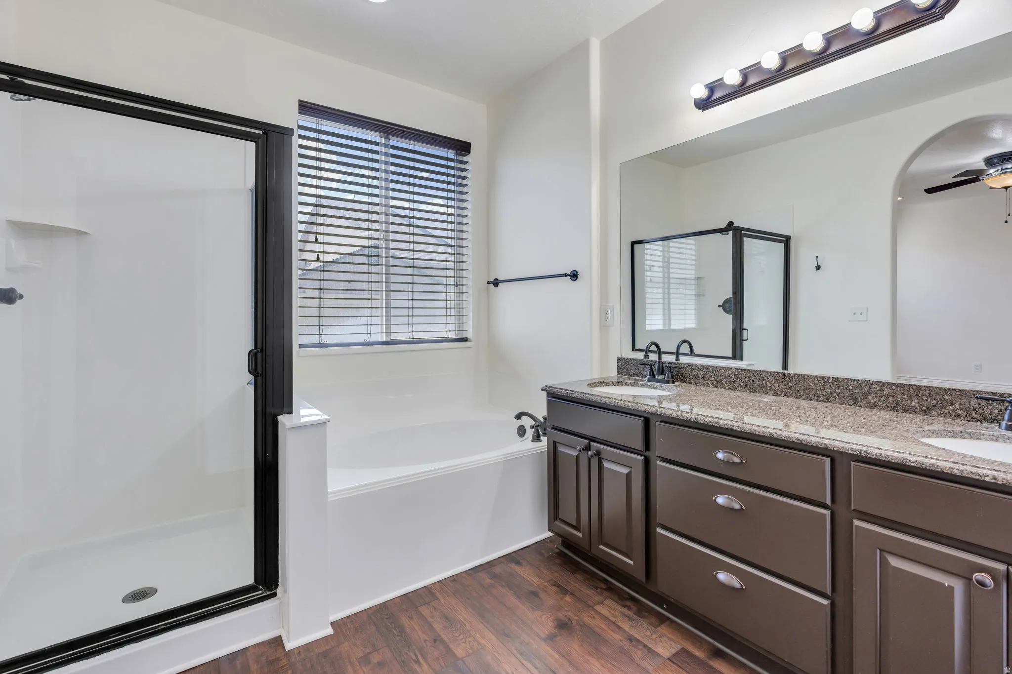 Primary bath with dual vanity, undermount sinks, and dark cabinetry, large mirror, wood-finish flooring, separate shower with dark-framed enclosure, and integrated bathtub