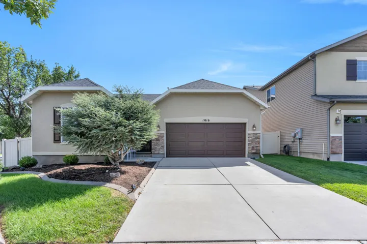 Stucco exterior with stone accents, attached two-car garage, concrete driveway, front landscaping with mature tree, and white fencing