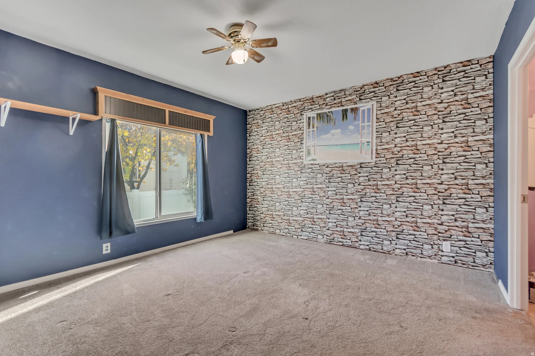 Carpeted room featuring a ceiling fan with light fixture, a window with a wood-finish valance, and an accent wall with a stone-finish