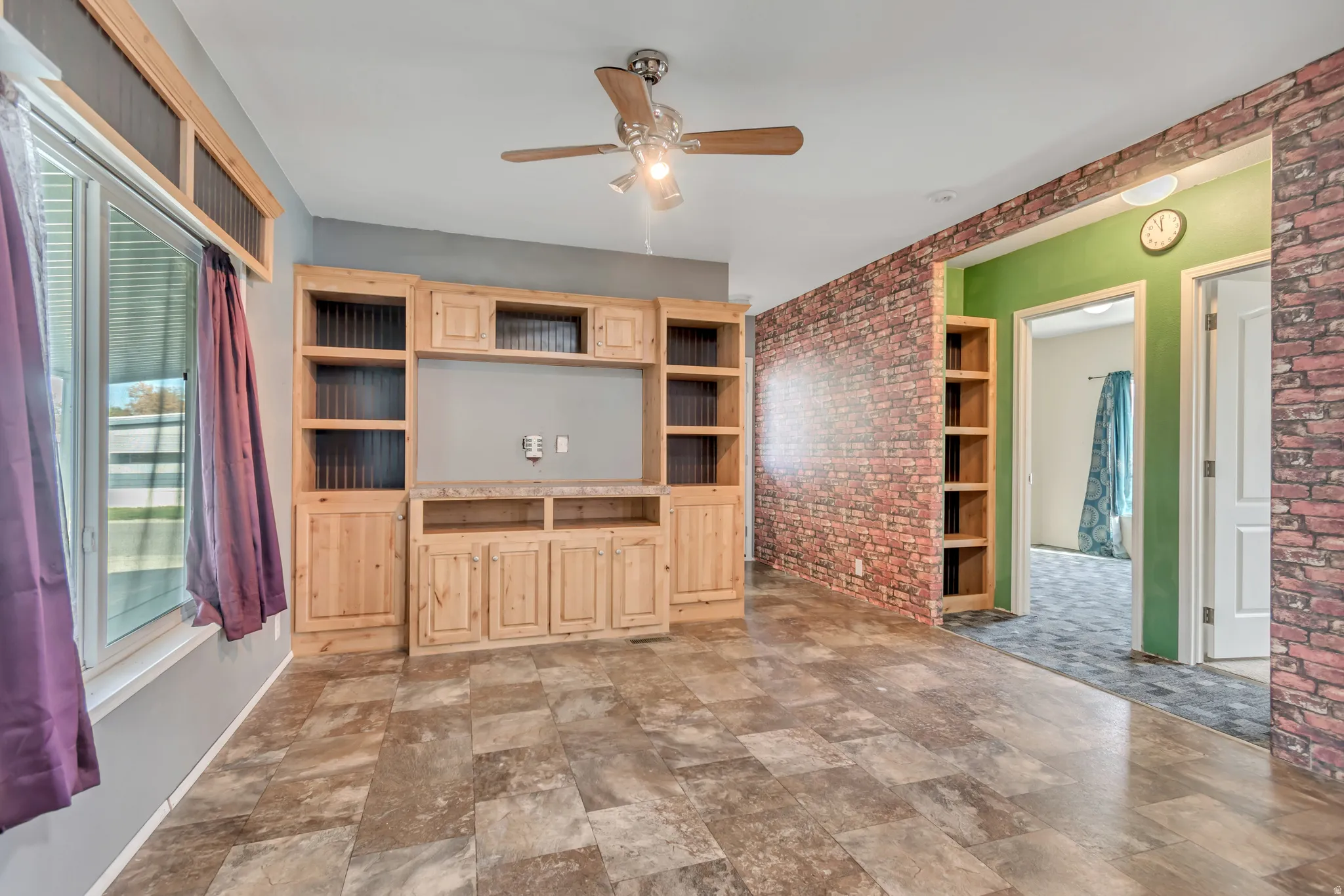 Built-in entertainment center with natural wood-finish cabinetry, tile-look flooring, ceiling fan, and exposed brick-look accent wall