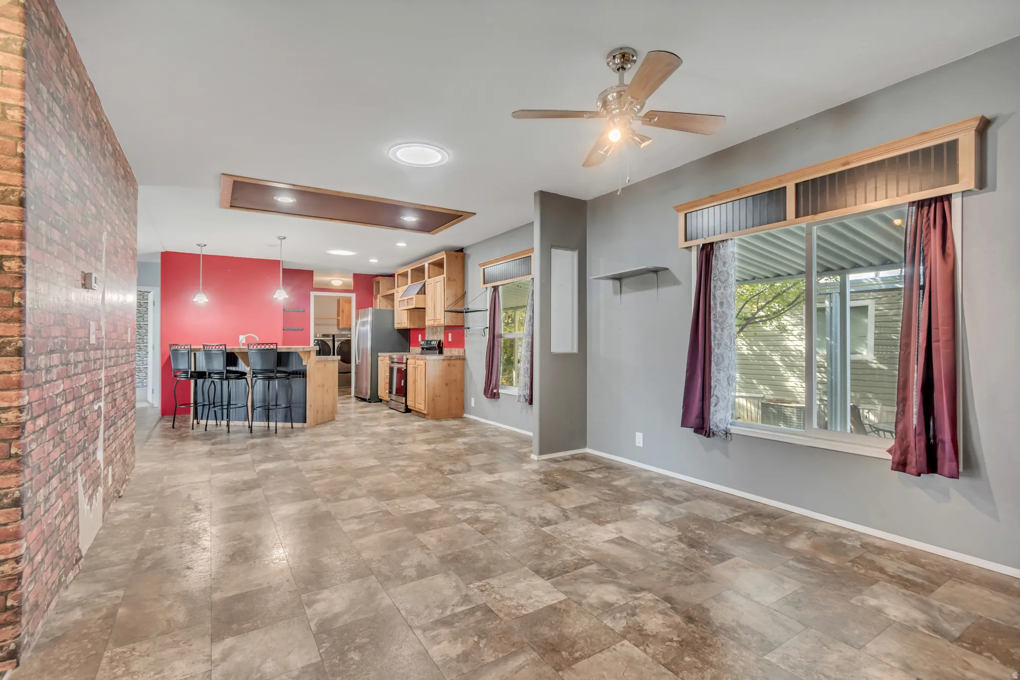 Open-concept living area with tile flooring, exposed brick wall accent, kitchen island with pendant lighting, wood cabinetry, and a ceiling fan