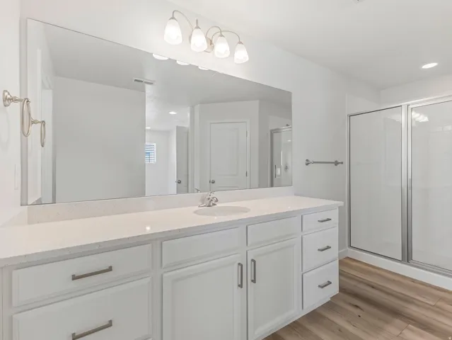 White vanity with dual sinks, a stone-finish countertop, brushed nickel hardware, and wood-finish flooring