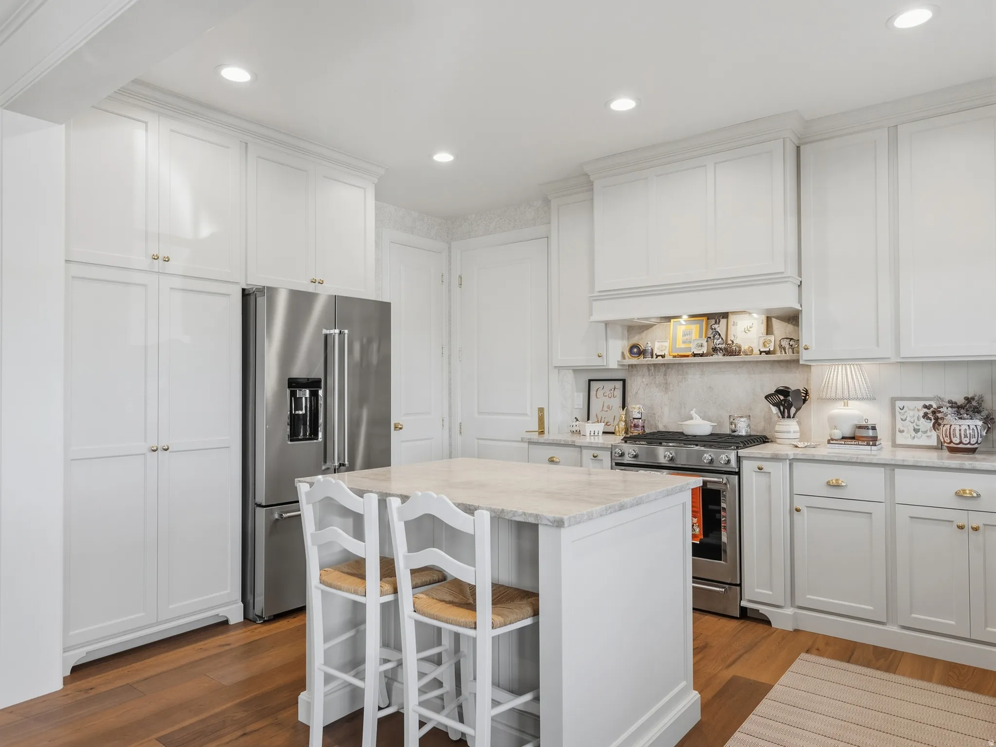 Kitchen featuring white cabinetry with gold-tone hardware, wood-finish flooring, a central island with a light-toned countertop, a stainless steel refrigerator, and a stainless steel range with a built-in hood