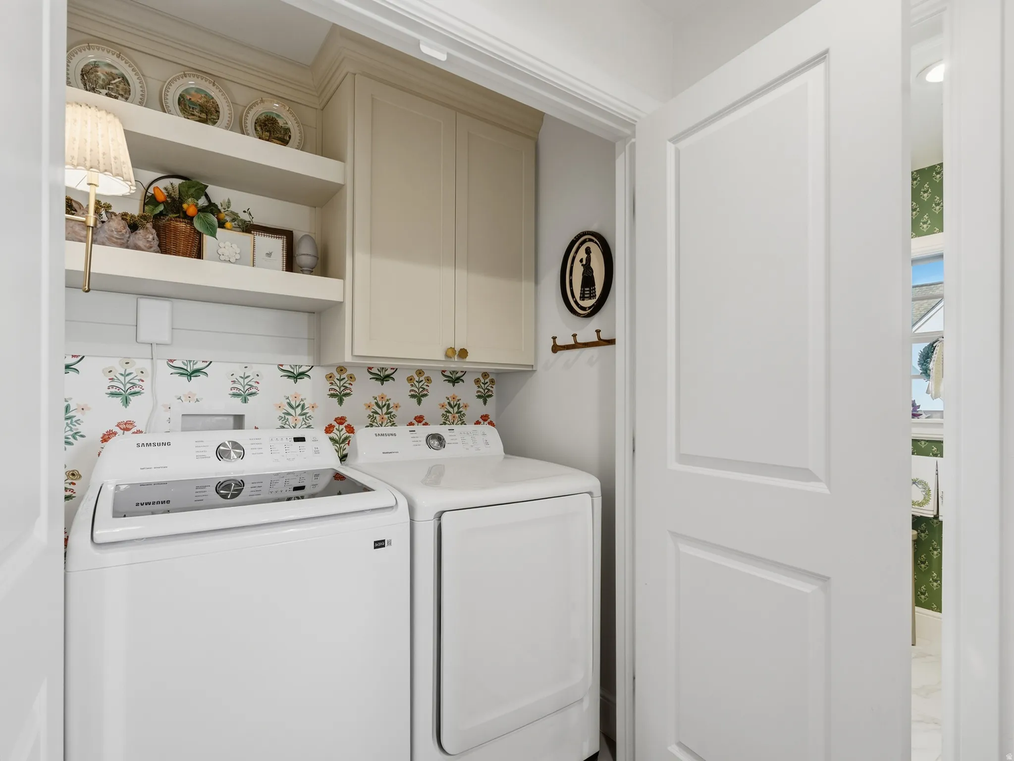 Dedicated laundry room featuring white front-load washer and electric dryer, decorative tile backsplash with botanical motif, custom cabinetry with upper shelving for storage, and recessed lighting throughout
