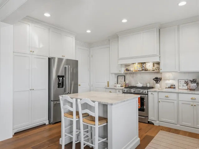 Kitchen featuring white cabinetry with gold-tone hardware, wood-finish flooring, a central island with a light-toned countertop, a stainless steel refrigerator, and a stainless steel range with a built-in hood