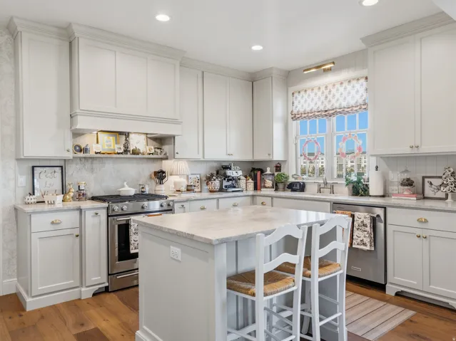 White shaker-style cabinetry, stainless steel appliances, kitchen island with stone countertop, wood-finish flooring, and a large window above the sink