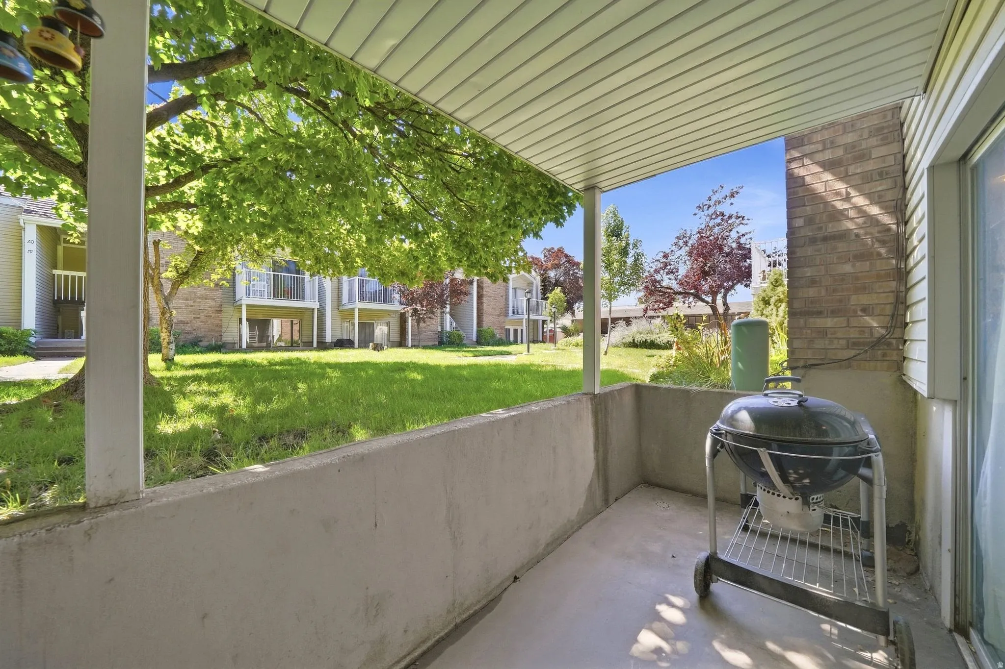 Covered patio with concrete flooring, brick exterior wall, and direct access to a large communal lawn featuring mature trees