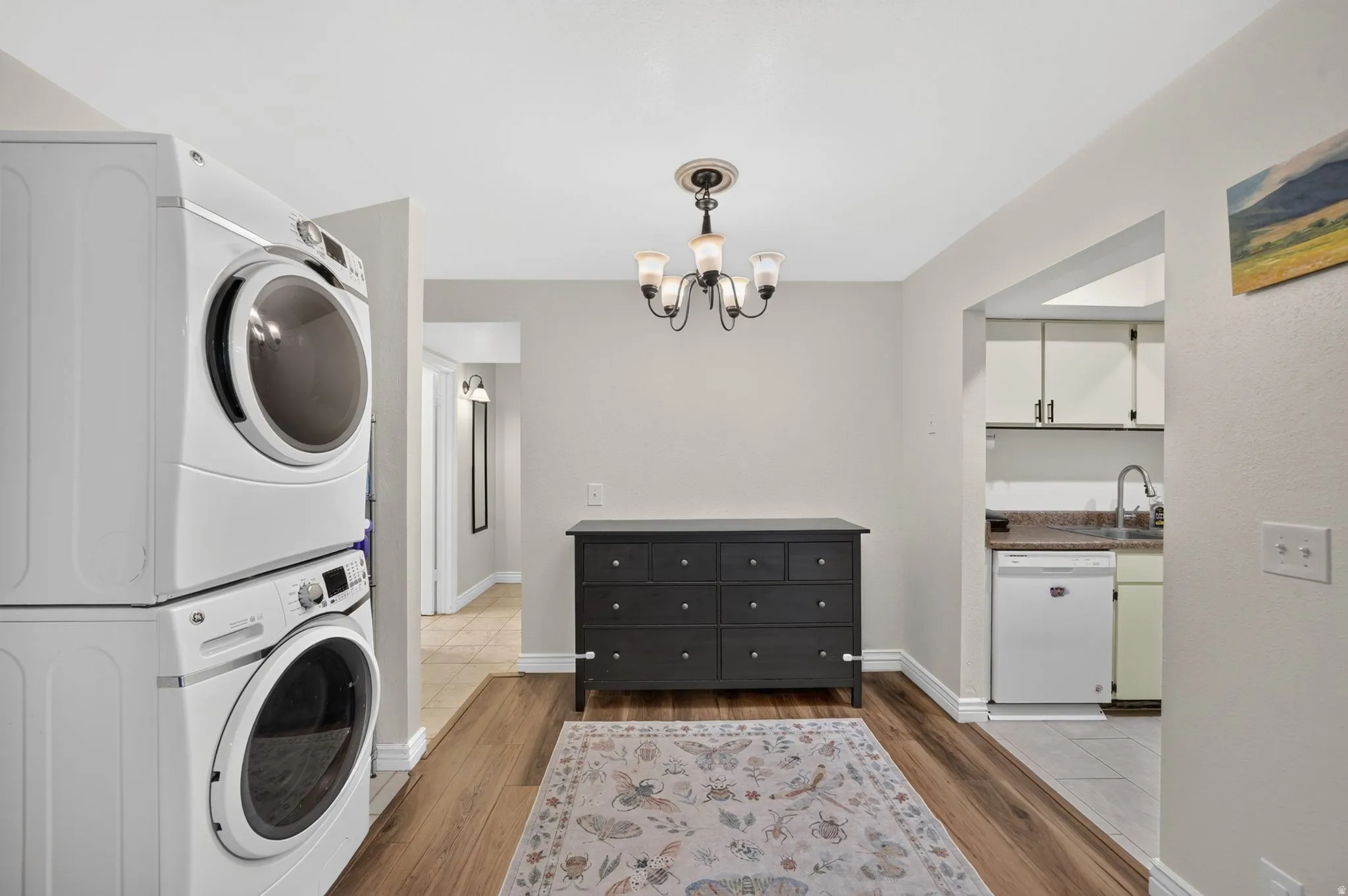Stackable front-load washer and dryer, wood-finish flooring, and an adjacent kitchen with white cabinetry and a dishwasher