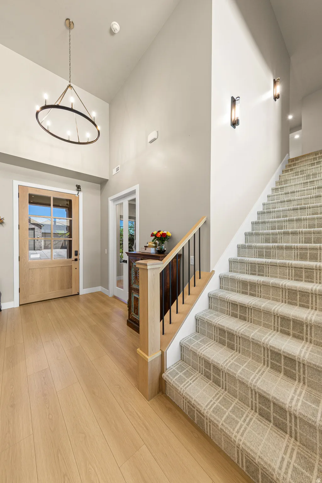 Light wood-finish flooring with a multi-paned wood entry door, high ceilings, and a staircase with plaid carpet and wood railing