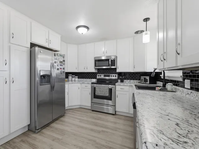 White shaker-style cabinetry, stainless steel appliances, dark tile backsplash, light wood-finish flooring, and white stone countertops