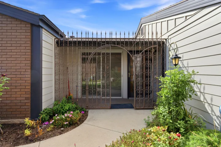 Wrought iron security gate entry with arched details, brick exterior accents, and a concrete pathway flanked by garden beds