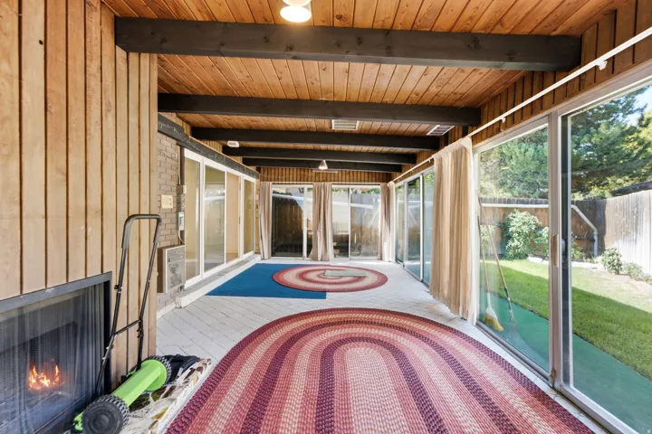 Enclosed patio with wood-paneled walls and ceiling, exposed beams, a built-in fireplace, and sliding glass doors opening to a grassy yard