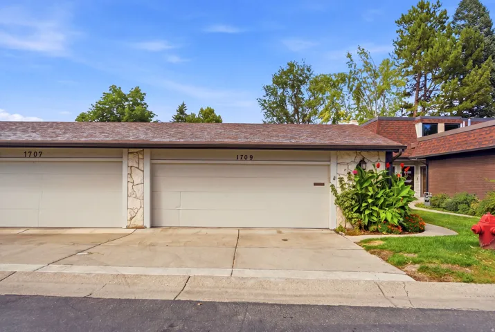 Dual garage doors with stone veneer detailing, a shingled roof, mature landscaping, and a brick exterior element