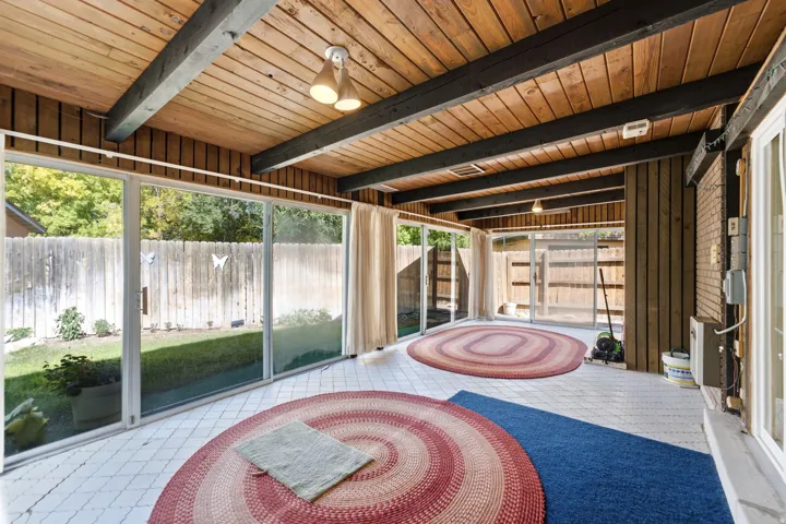 Wood-paneled ceiling with exposed beams, multiple sliding glass doors, white tiled flooring, and a brick accent wall