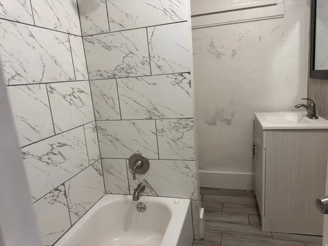Bathtub with tile surround, wood-finish flooring, and a single vanity with integrated sink