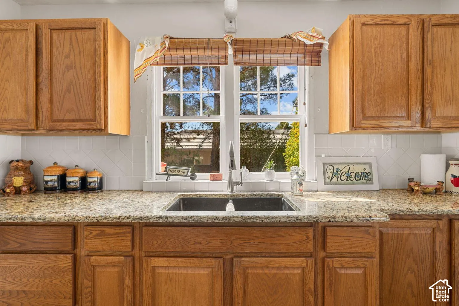 Wood cabinetry with panel doors, stone-finish countertops, stainless steel undermount sink with a gooseneck faucet, white tile backsplash, and a double-hung window