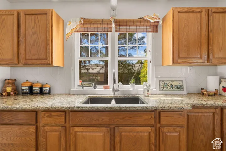 Wood cabinetry with panel doors, stone-finish countertops, stainless steel undermount sink with a gooseneck faucet, white tile backsplash, and a double-hung window
