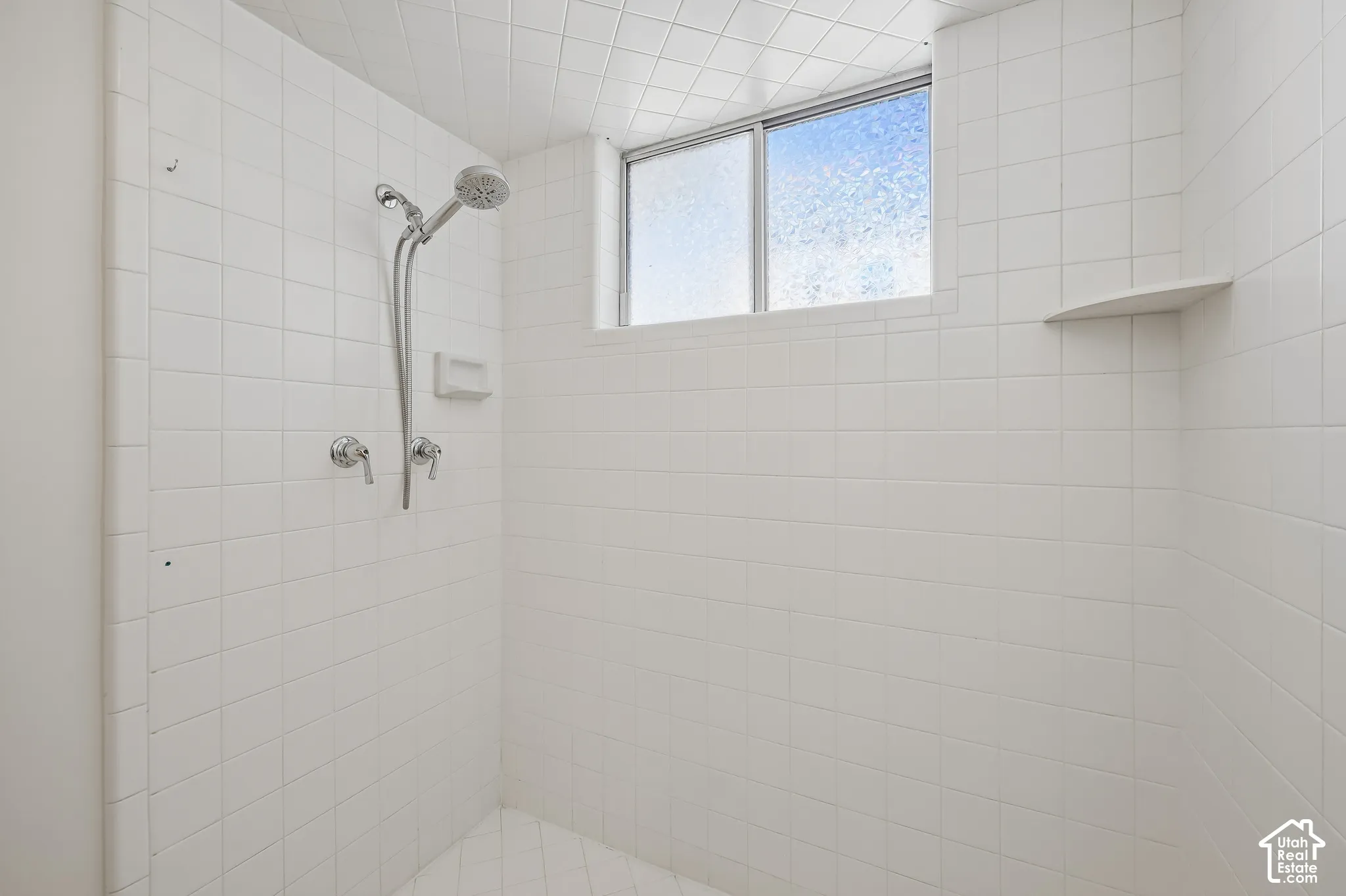 Tiled shower stall featuring a chrome-finish shower head with handheld sprayer, two chrome-finish control knobs, a built-in soap dish, a frosted window, and a corner shelf