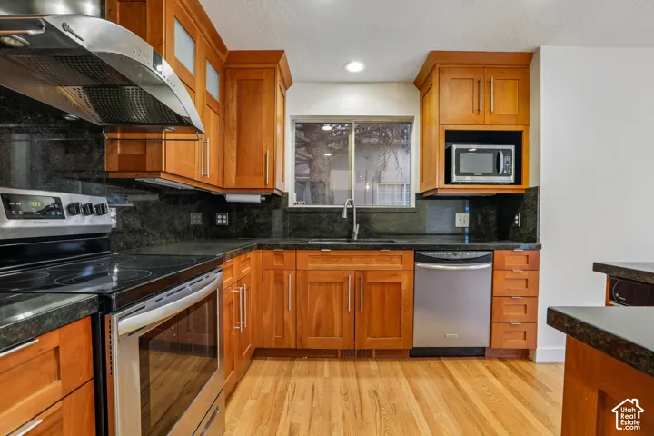 Wood cabinetry with stainless steel hardware, dark stone countertops, wood-finish flooring, and built-in stainless steel appliances