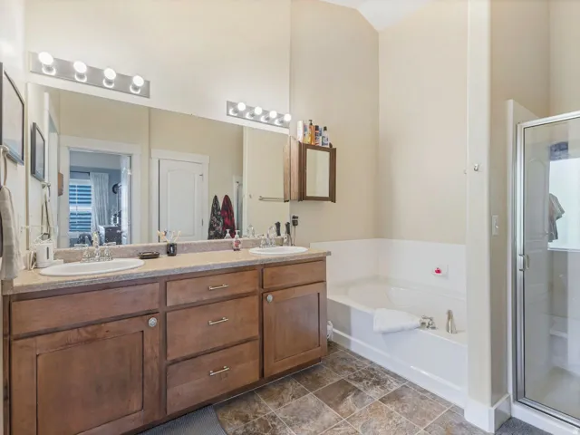 Dual vanity with integrated sinks, wood-finish cabinetry, built-in soaking tub, separate glass-enclosed shower, and tile flooring