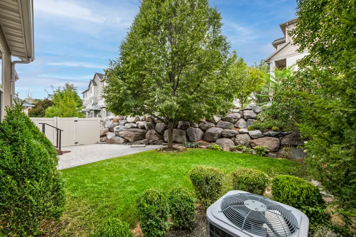 Expansive grass lawn with mature trees and established shrubbery, featuring a stone retaining wall and paver patio area