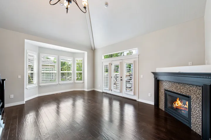 Vaulted ceiling, dark wood-finish flooring, bay window with plantation shutters, fireplace with mosaic tile surround, and French doors with transom windows
