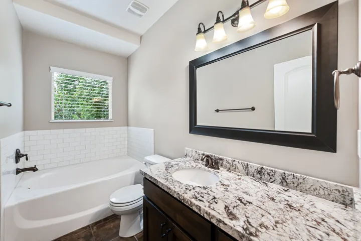 Bathroom featuring a built-in tub with subway tile surround, dark wood-finish vanity with stone countertop, undermount sink, and a framed mirror with a four-light fixture